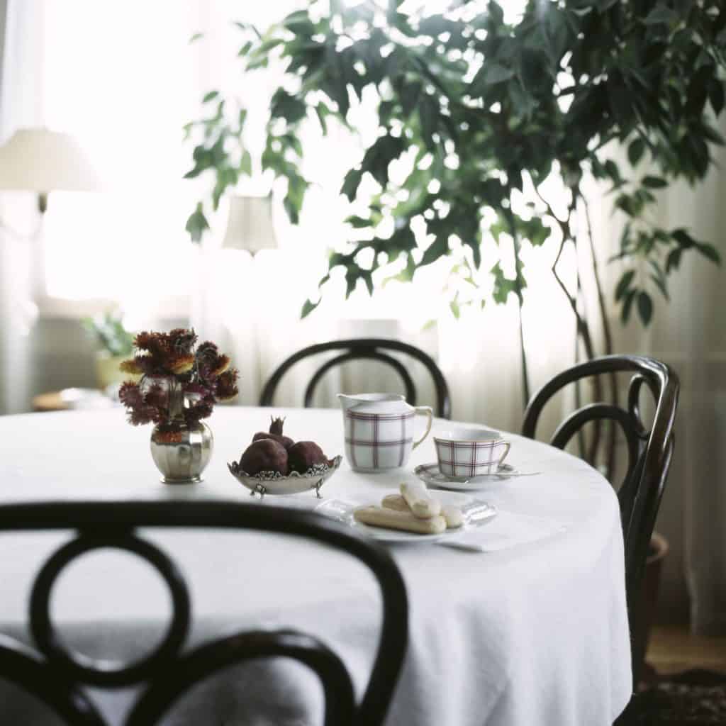 A white table cloth on a round dining table with tea cups, coasters, and a small indoor plant