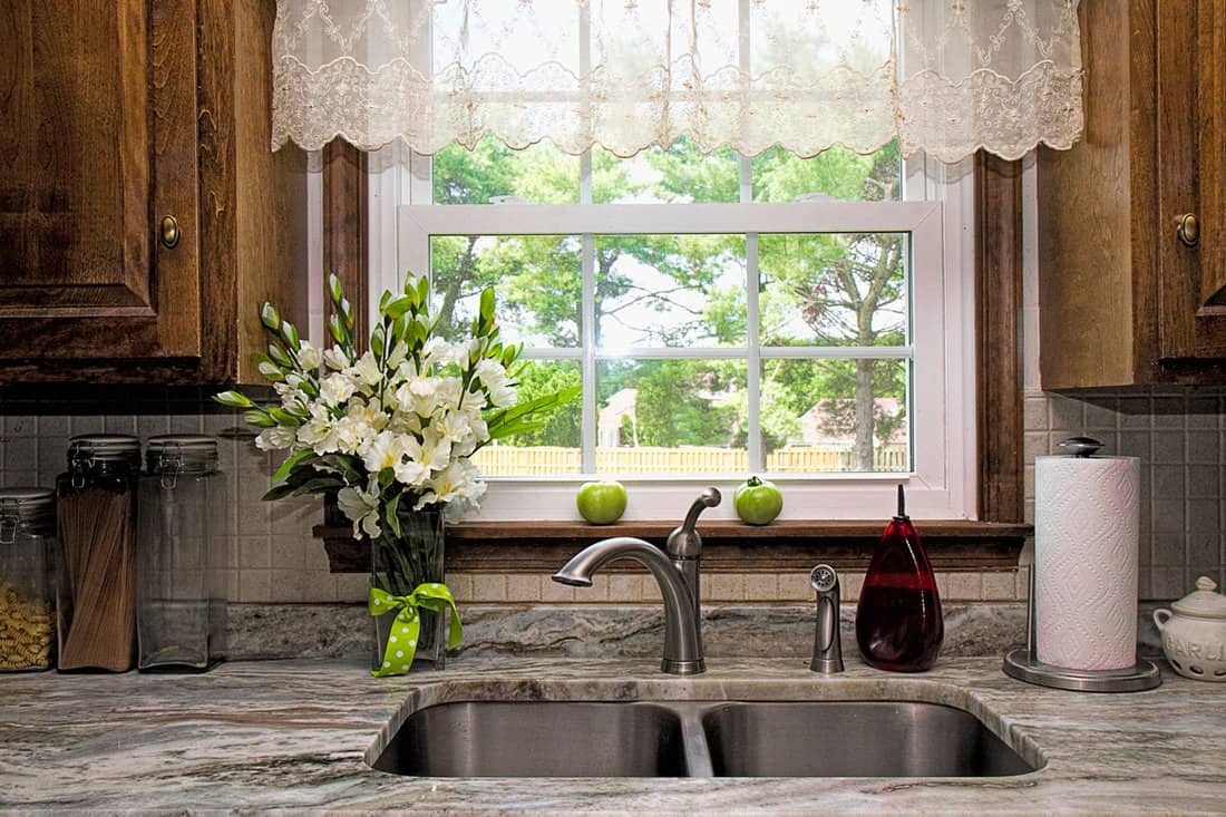 View of Kitchen sink, window view of backyard, curtains and flowers. Two green tomatoes on window seal witing to ripe. Beautiful designer gray-white-green granite countertop.