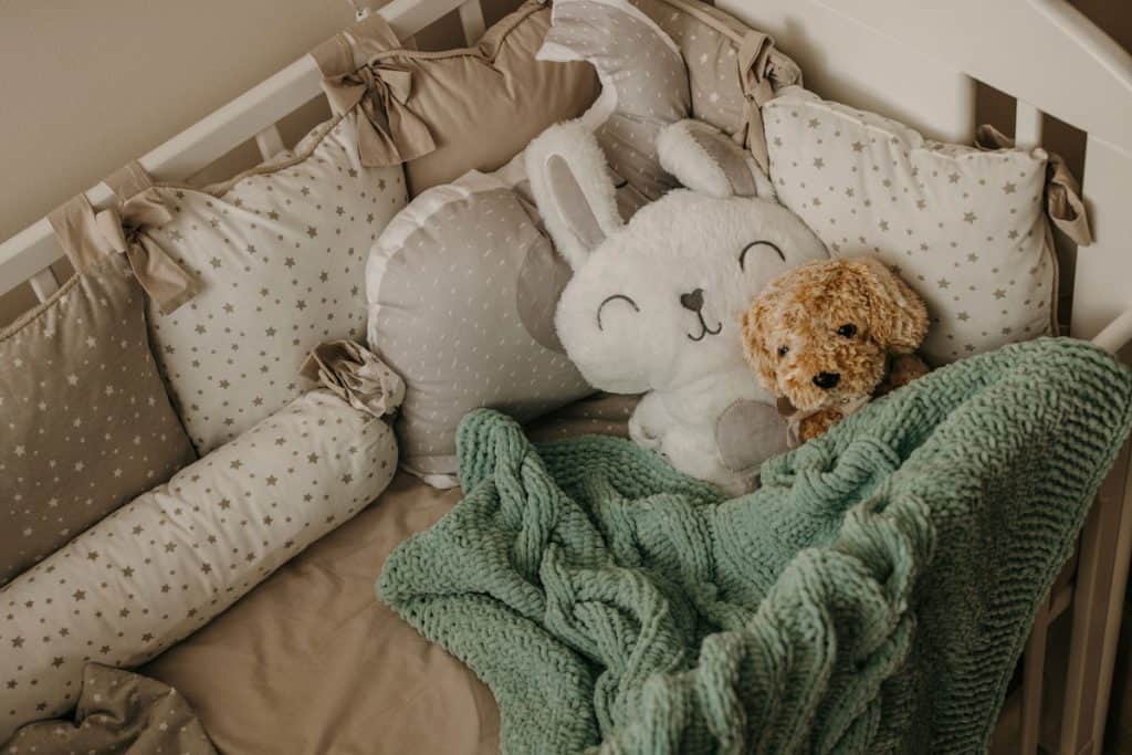 A small white teddy bear placed on a babies crib with lots of pillows and a green blanket