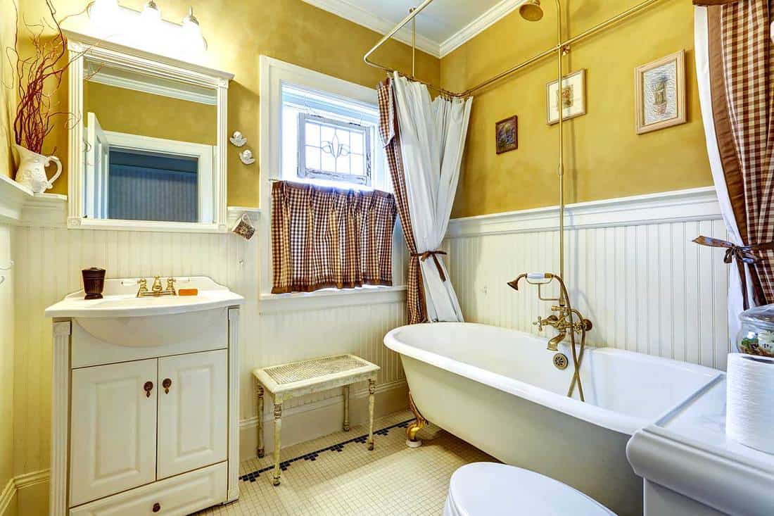 Yellow old bathroom interior with white plank paneled wall trim and antique vanity and bath tub