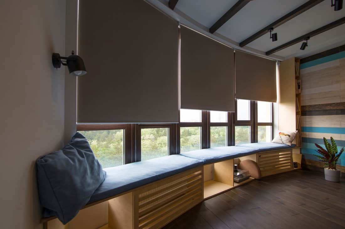 Motorized roller shades in the interior. Automatic roller blinds beige color on big glass windows. Remote Control Shades are above the windosill with pillows. Summer. Green trees outside.
