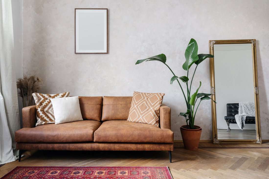Modern design of living room with brown eco leather couch, soft cushions, mirror with golden frame, copy space picture frame on wall and houseplant in pot