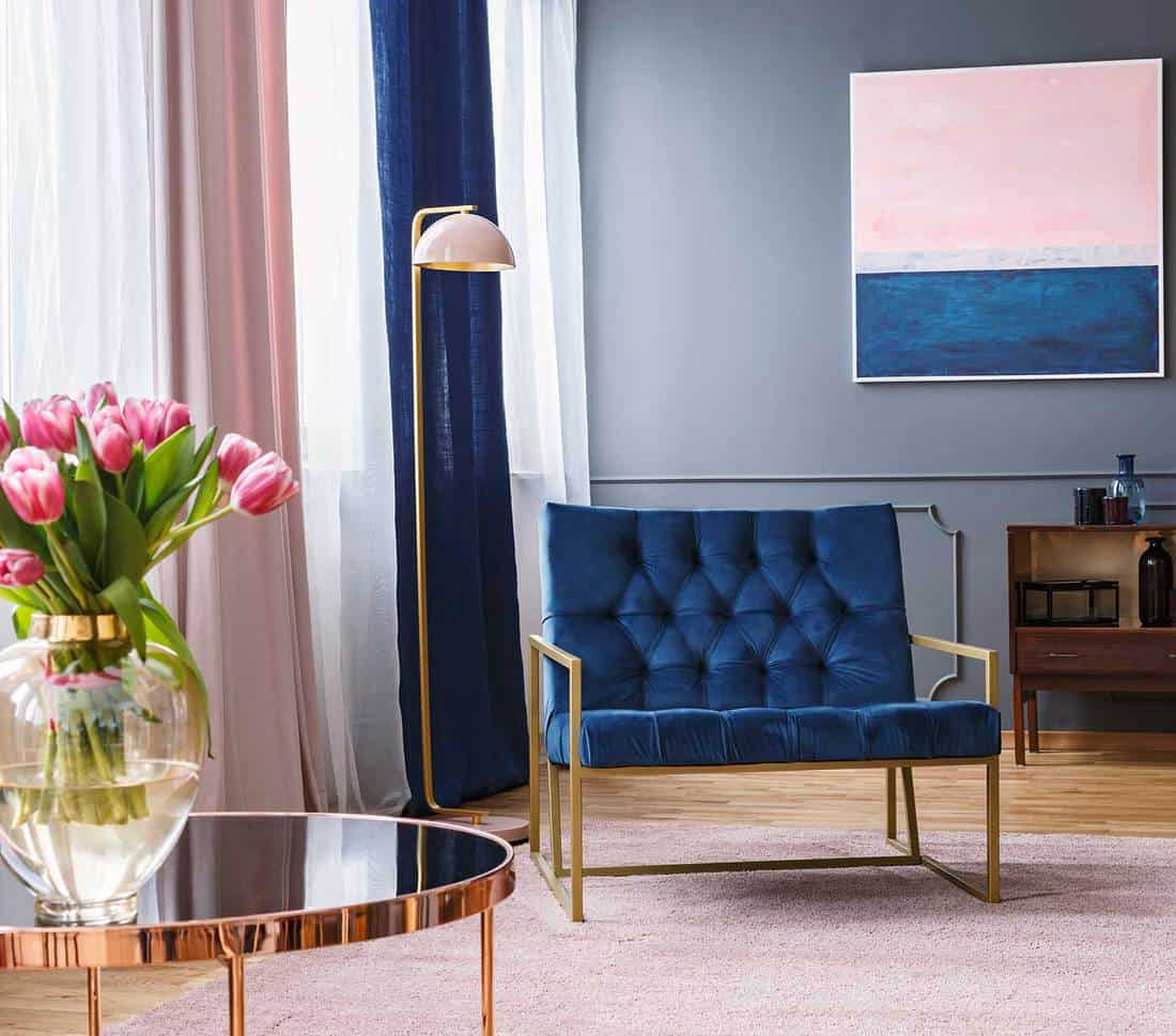 Navy blue armchair next to lamp in sophisticated apartment interior with painting and flowers