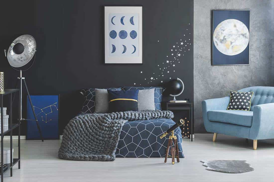 Telescope near bed with knit blanket against dark wall with star stickers in blue bedroom interior