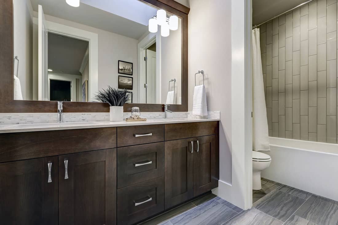White and brown bathroom boasts a nook filled with double vanity cabinet topped with white counter paired with white and grey tile backsplash under a framed mirror lit by sconces.