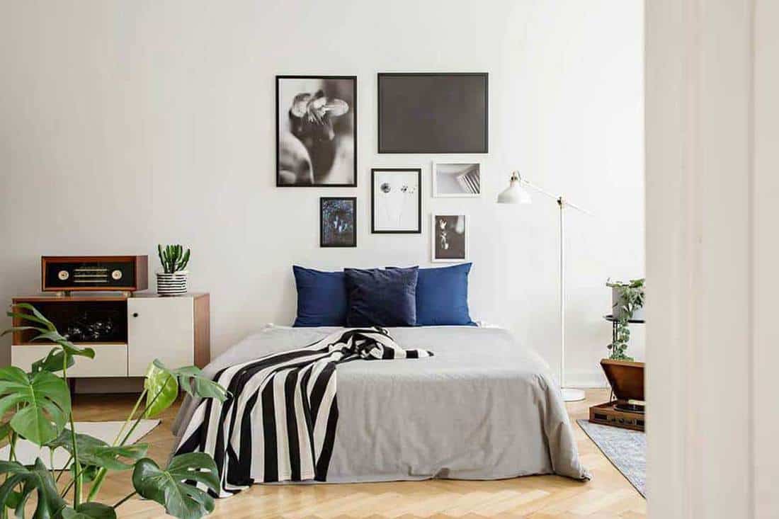 White wooden commode next to bed with dark bluepillows grey duvet and striped black and white blanket in bedroom with framed art gallery on the wall