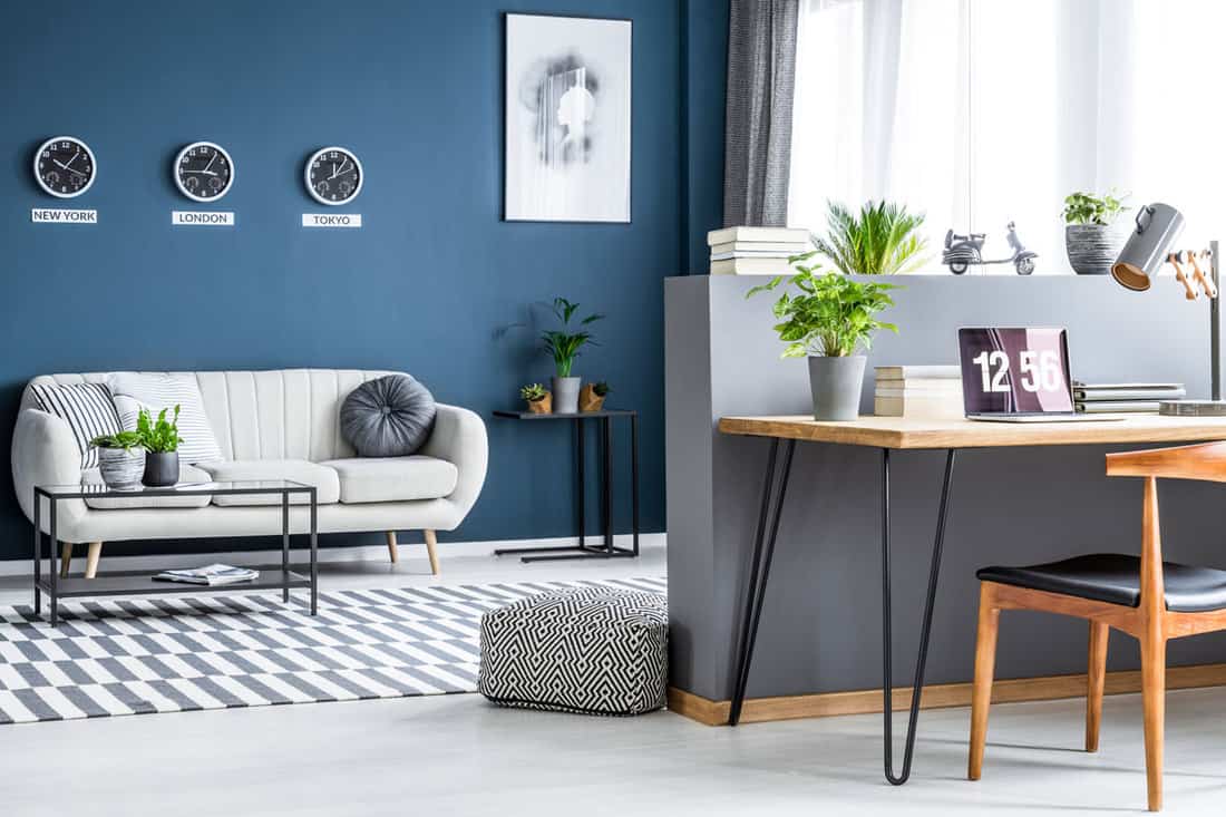 Donkerblauw woonkamerinterieur met drie klokken, eenvoudige poster, lichte bank en thuiskantoorhoek met laptop op haarspeldbureau