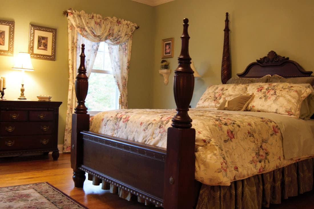 Victorian-style bedroom with a four poster bed, side table, windows, and curtains