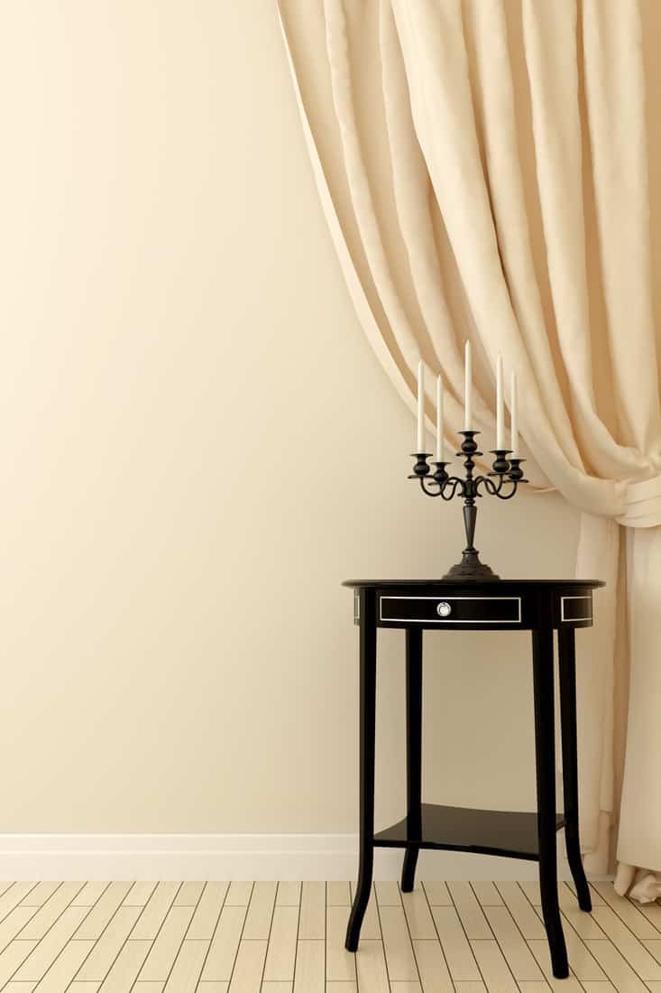 Beige colored wall with light yellow colored curtains and unlit candle on top of black wooden table