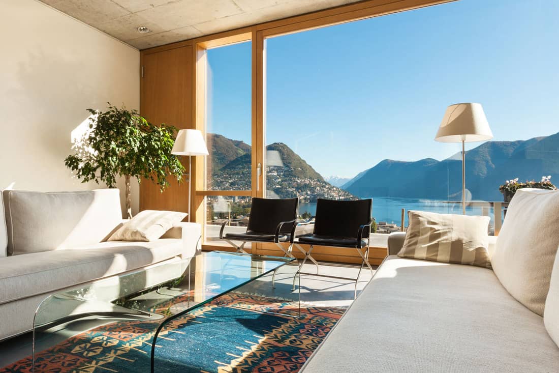 Modern living room with dirty white colored sofa placed near huge glass windows showcasing a beautiful mountainous scenery.