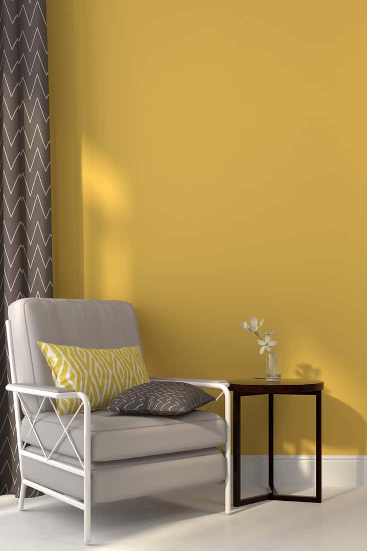 White armchair and a dark wooden table against a background yellow wall with complementing patterned curtains and throw pillows