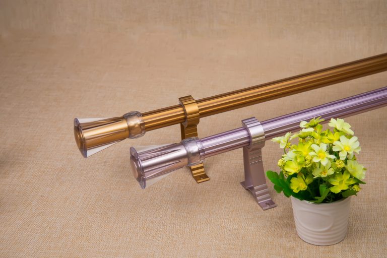 Brass and silver curtain rods placed on the floor with a small flower vase on the side, How To Choose Curtain Rods [You MUST check these 3 things]