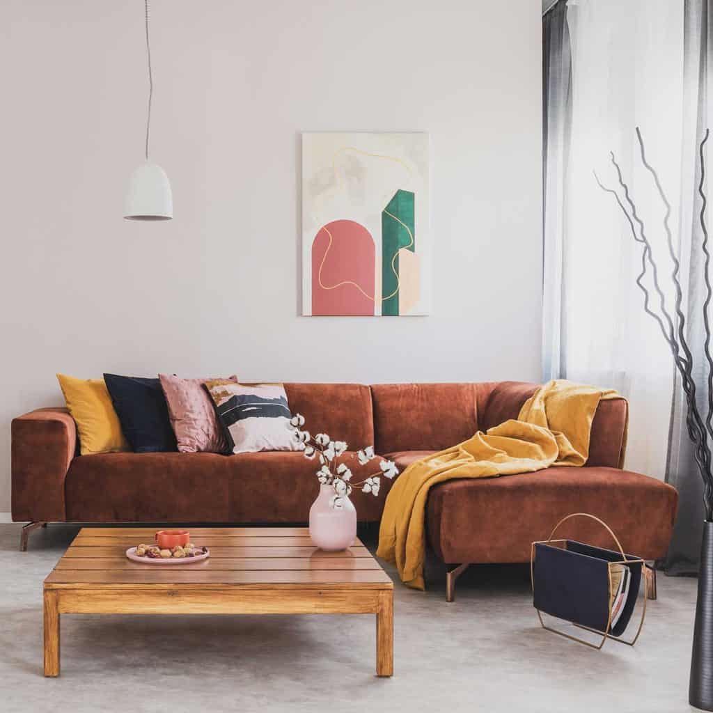Flowers in vase on wooden coffee table in fashionable living room interior with brown corner sofa with pillows and abstract painting on the wall
