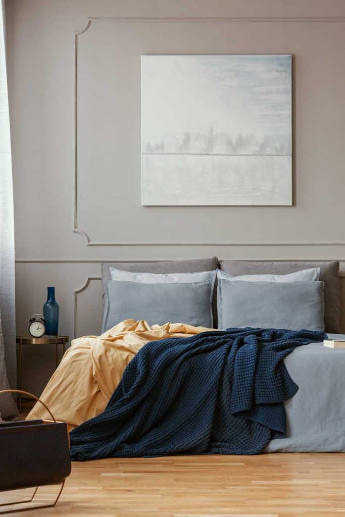Trendy bedroom interior with painting on gray wall, cozy bed, bedside table with alarm clock