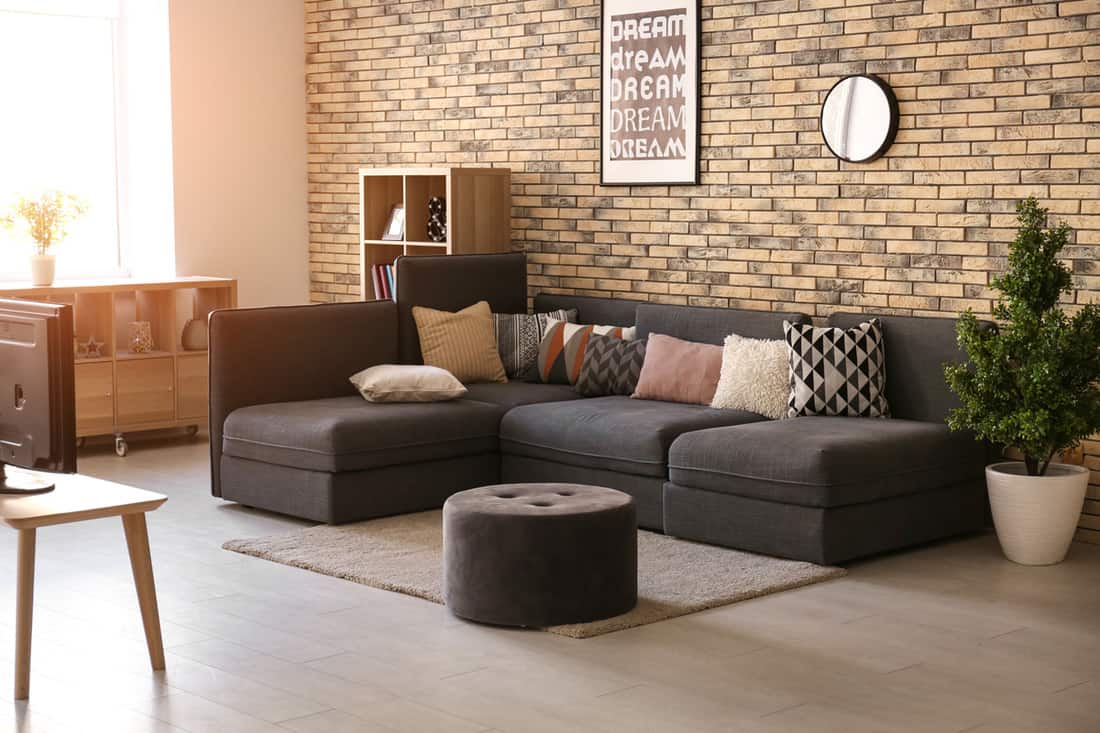 A modern living room with a dark colored sectional sofa and an ottoman up front for a foot rest, Can An Ottoman Be Used For Sitting?