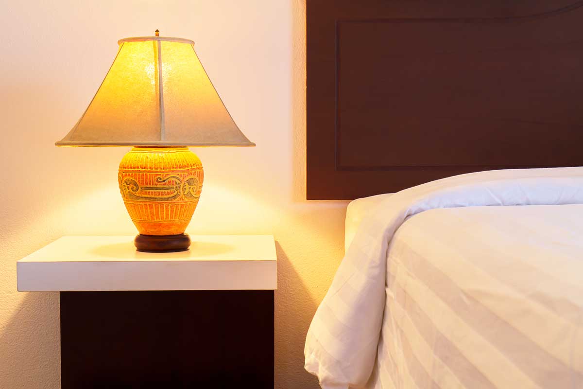 Lamp on a night table with light switched on beside the bed in a cozy bedroom, How To Decorate A Lampshade [Best 4 Suggestions]