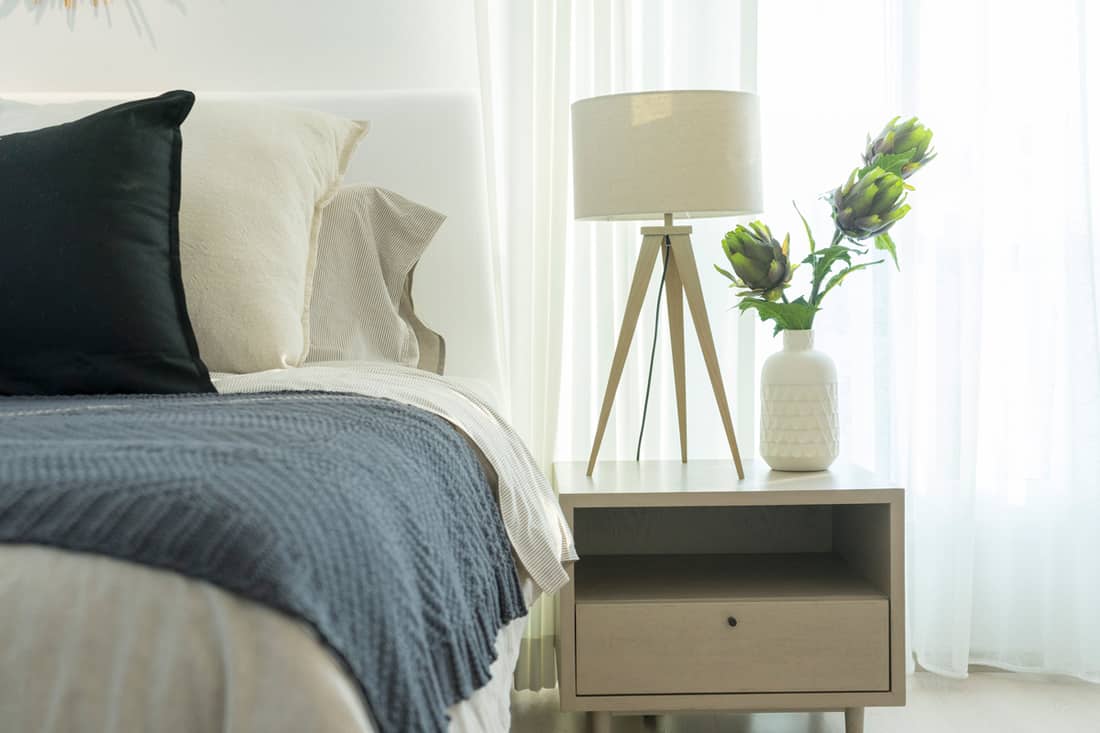 A cozy white bedroom with a box shaped nightstand and lampshade placed on top, Should Nightstands Be Taller Than the Bed?