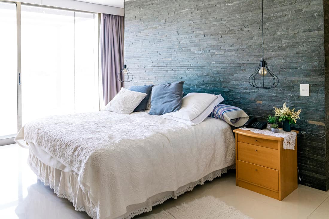 A modern bedroom with a white laced blanket, blue throw pillows, faux stone decorated header wall, and a brown colored nightstand, Does Nightstand Have to Match Dresser? [Bedroom Furniture Coordination Tips]