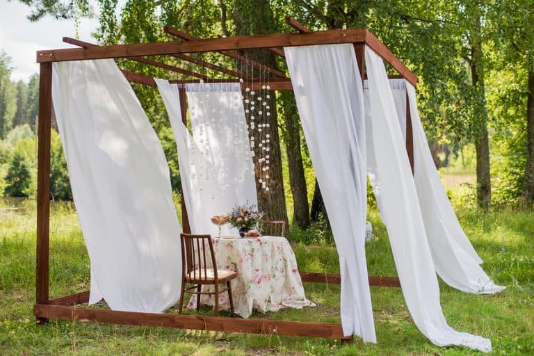 An outdoor gazeebo with white outdoor curtains and a dining table with chairs in the middle, How To Keep Outdoor Curtains From Blowing