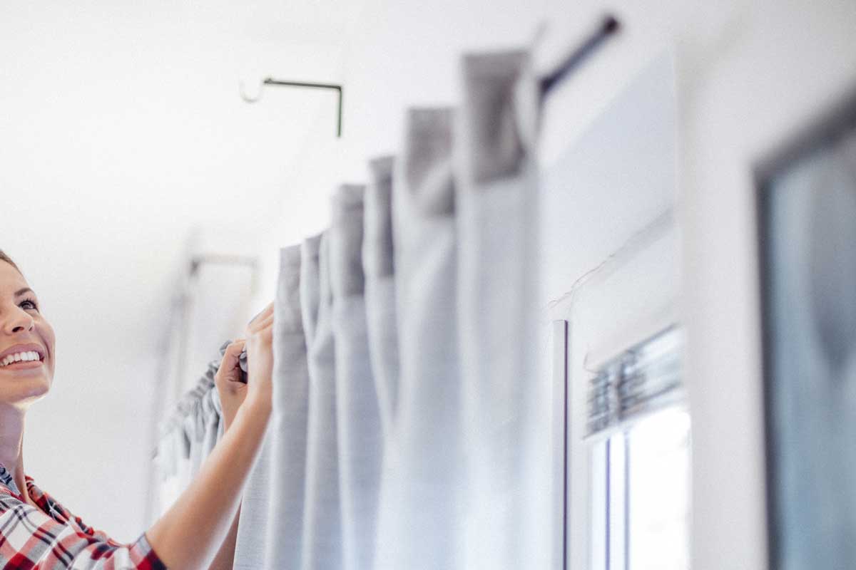 Beautiful Caucasian smiling woman in casual clothes is hanging curtains after washing at cozy home, 4 Easy Ways to Make Curtains Slide Easily