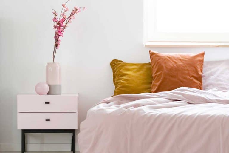 Feminine bedroom interior with pink sheets on a bed standing near wooden bedside table, How To Decorate The Top Of A Nightstand Or Bedside Table? [7 Suggestions]