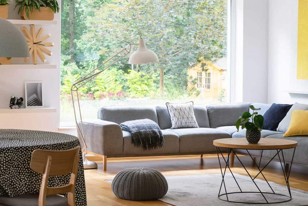 Pouf next to table in modern living room interior with gray corner sofa and window
