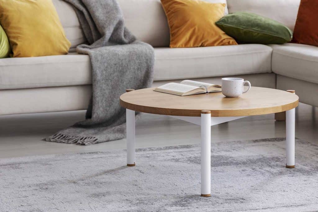 Round wooden table with book and cup on gay carpet next to sofa in flat interior