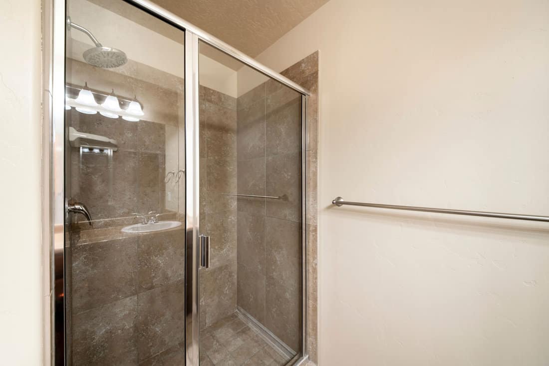 Shower stall inside a bathroom with glass panel and metal frames. There is a metal handrail on the wall near the closed shower stall with corner shelf on the tile wall surround.