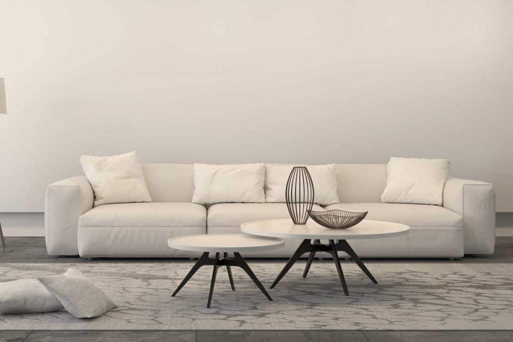 White sofa with set of coffee tables, a floor lamp and pillows. Interior is white, brightly lit with ornate carpet. Concrete industrial floor and a white wall behing