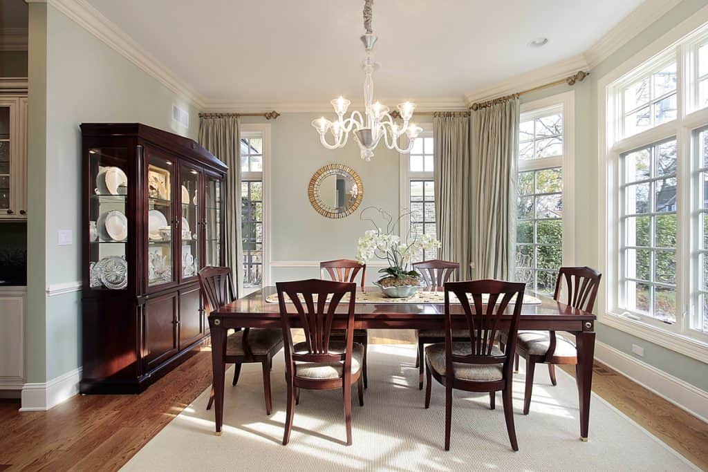 Een klassieke eetkamer met een koloniaal thema met houten stoelen en een tafel gecombineerd met een wit tapijt onder de tafel en een klassieke kast aan de linkerkant