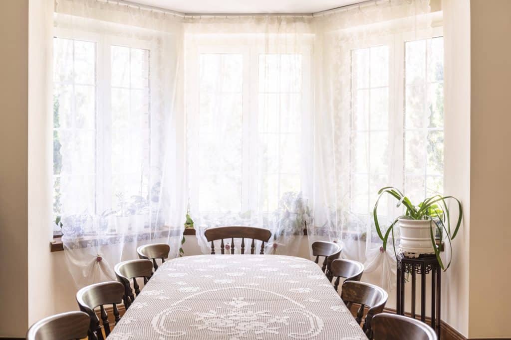 A dining area lit by a huge bay window with white flora curtains