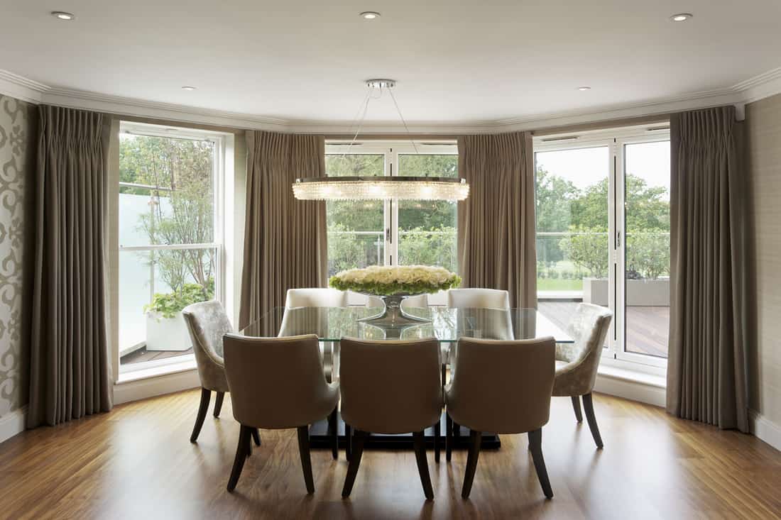A dining are with brown classic designed dining chairs and a bay window with cream colored curtains, What Curtains Look Best On Bay Windows?