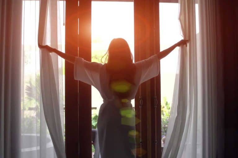 Young woman in bathrobe open curtains and stretch standing near the window at home, Do Curtains Stay When Selling A House?