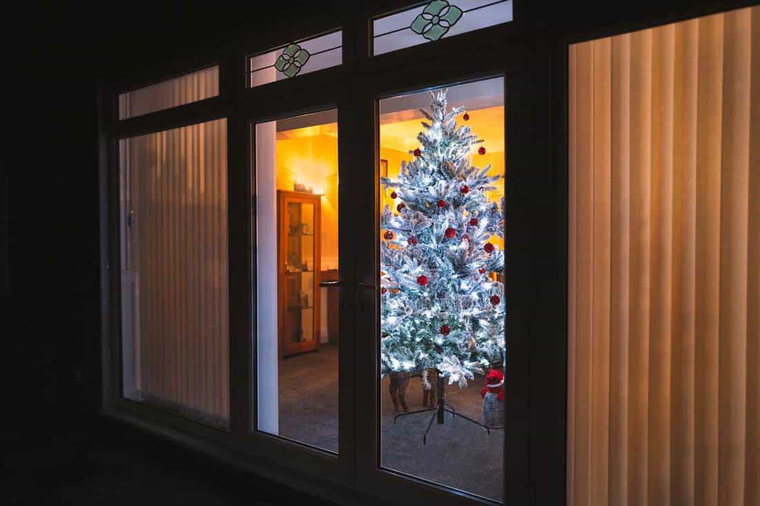 A frosted white artificial christmas tree with red and silver decorations seen through large french doors with vertical slat blinds, Can You Put Blinds On French Doors? (And How To)