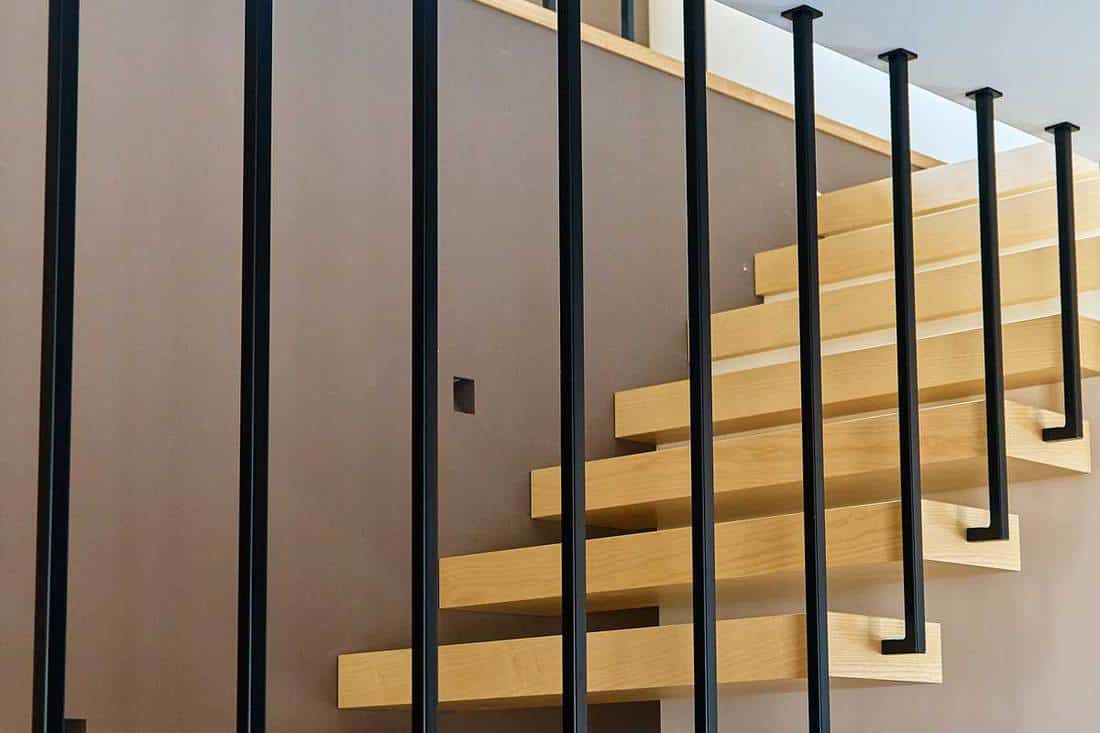 Ash lumber stairs with railing attached to brown wall and leading to second floor inside stylish modern house, How Much Do Floating Stairs Cost? [Here's the Breakdown]