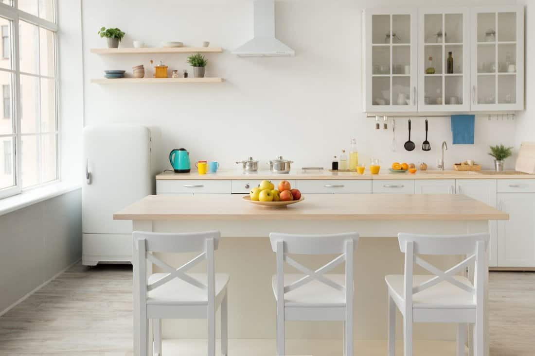 Dining room, kitchen, healthy food and stylish design. Bright apples on plate on table, white chairs, blue kettle, colored cups and utensils on furniture, small refrigerator, range hood, light walls