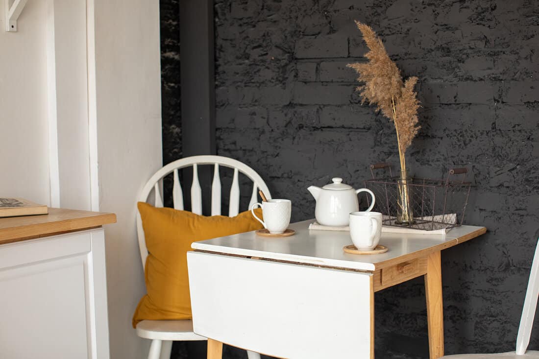 Folding table with cups and white tea pot and chair with pillow against black brick wall in kitchen