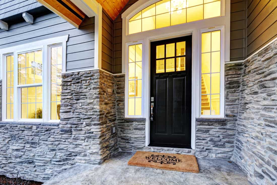 A modern colonial house with decorative stones on the porch and a black door with framing painted in white, How To Hang A Door In An Existing Frame? [7 Steps]