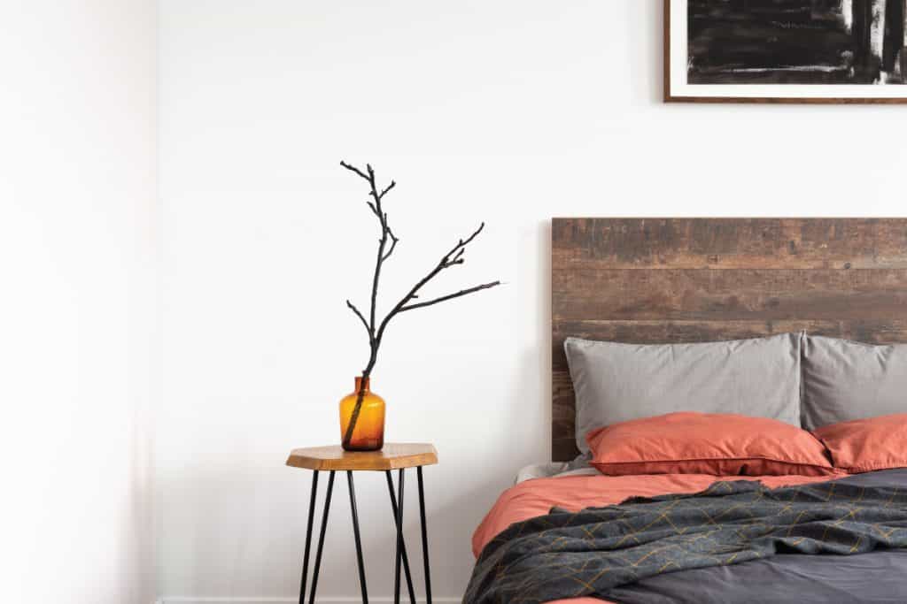 Orange glass vase with a tree branch standing on the wooden table next to bed with colorful sheets