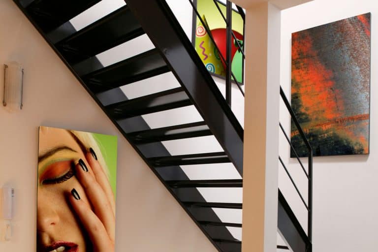 A residential steel staircase with hanging paintings on the wall, How to Hang Art on Staircase Wall [Inc. Picture Arrangement Tips]