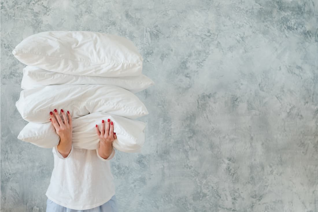 Woman holding big pile of white soft cozy pillows on gray background, Should New Pillows Be Washed Before Use?
