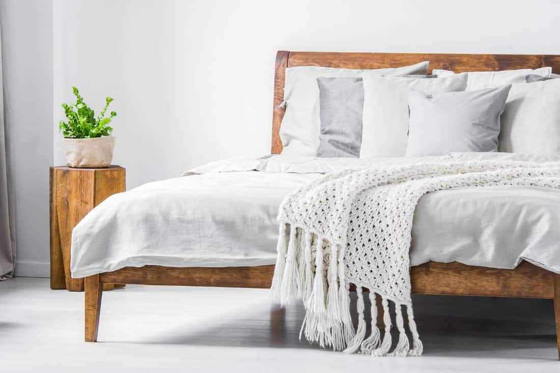 Wooden framed comfortable bed with many pillows, blanket and sheets and a sideboard with flowers on top in a white stylish bedroom interior.