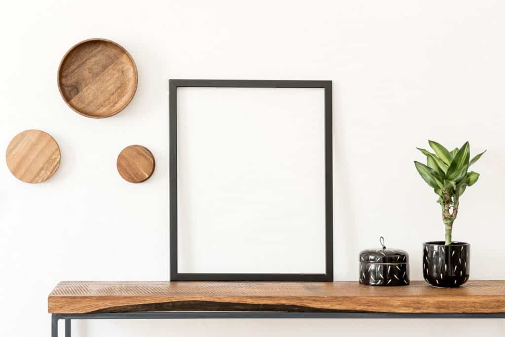 A black mock up picture frame placed on top of a wooden table with a small table and plant on the side