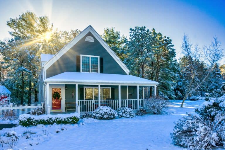 Blue post colonial house with a roof and front yard covered in snow, How to Close Off the Porch for Winter (4 Porch Panel Ideas)