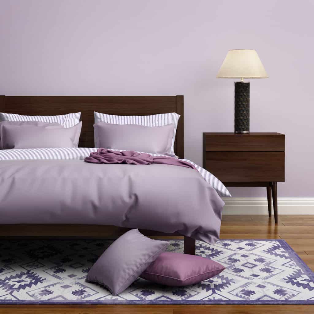 A brown bed with violet bedding, pillows, and area rug ow