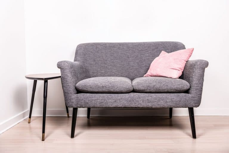 A gray loveseat with a pink throw pillow, and a small bar stool on the side inside a white living room, How Much Does a Loveseat Weigh?