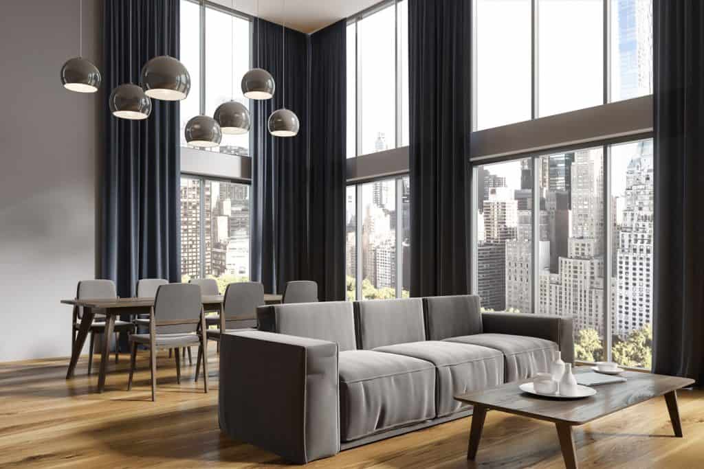 Un immense salon avec de hauts rideaux bleu foncé, d'immenses fenêtres, des meubles de couleur grise et un parquet en bois dur