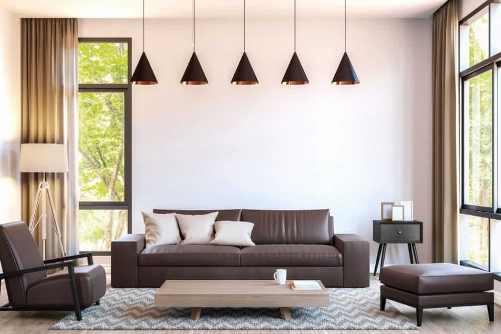 A luxurious living room with a white painted wall, huge sliding windows, cone shaped hanging lamps and brown couches with a zigzag patterned rug