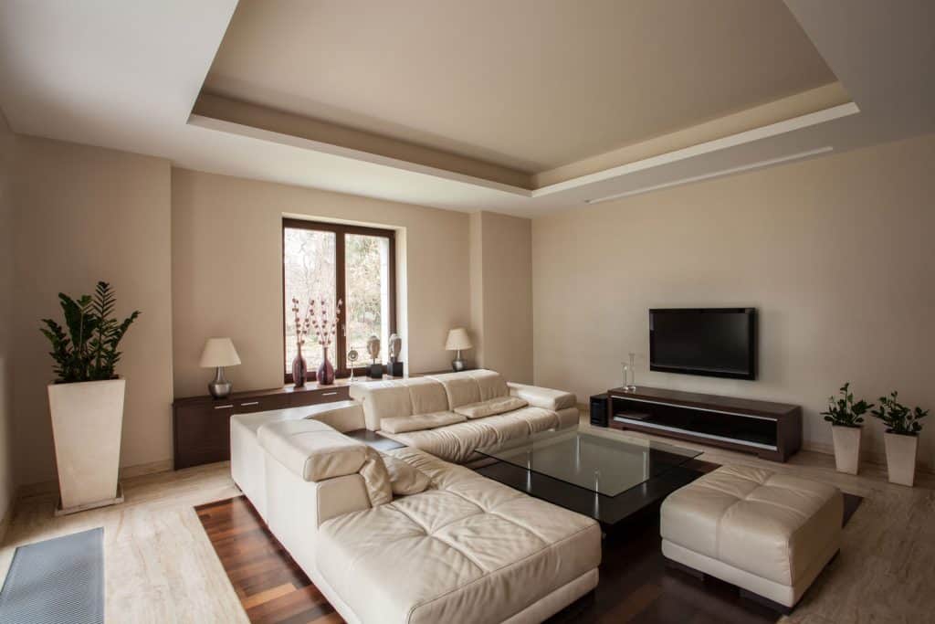 A modern living room with cream painted walls and decorated with a cream sectional couch, indoor plants and a brown wall unit with a wall mounted TV