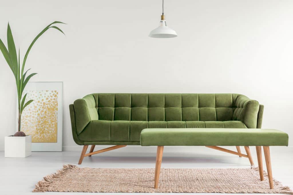 An olive green colored settee, small leather upholstered stool and a gray rug inside a white walled living room 